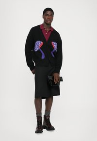 Cardigan negru cu motive colorate de flamingo, cămașă polo roșie, fustă neagră și cizme vișinii cu manșete tricotate gri. Poartă o geantă de mână.