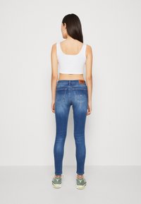 Jeans skinny azul oscuro con cintura alta, con bolsillos traseros y una etiqueta de cuero en la cinturilla. El modelo lleva una camiseta corta blanca y zapatillas.