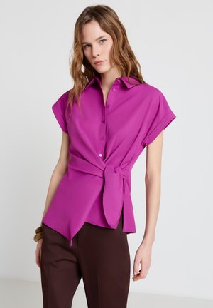 Femme aux cheveux châtain clair portant un chemisier magenta à manches courtes avec un nœud sur le côté et un pantalon marron foncé, debout devant un fond uni.