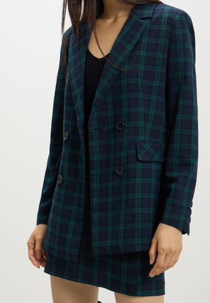 Femme portant un blazer croisé à carreaux vert foncé et bleu marine avec une jupe assortie, et un haut noir en dessous, debout sur un fond blanc.