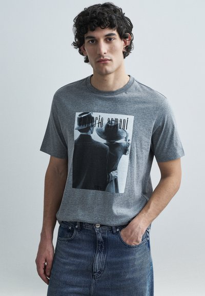 Grå bomuld T-shirt med sort-hvidt tryk af to figurer med hatte. Korte ærmer, rund hals, kombineret med blå denimjeans.