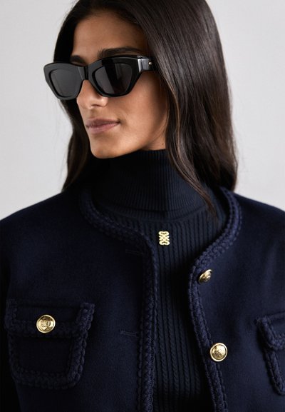 Sorte solbriller med et dristigt geometrisk design, parret med en marineblå struktureret rullekrave og en matchende jakke med guldknapper.