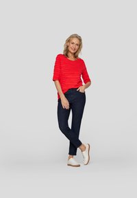 Roter strukturierter Oberteil mit Dreiviertelärmeln, kombiniert mit dunkelbeigen Slim-Fit-Jeans und weißen Schnürschuhen mit hellbrauner Sohle.