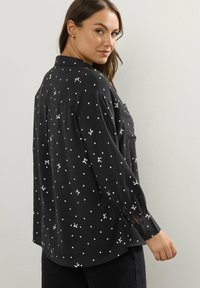 Maglietta nera a maniche lunghe con pois bianchi e piccoli disegni di fiocchi; presenta un colletto, polsini con bottone e un design dalla vestibilità rilassata.