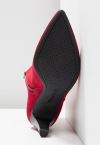 Botte à talon en suède rouge à bout pointu avec semelle noire texturée, fermeture éclair latérale et talon fin, vue du dessous.