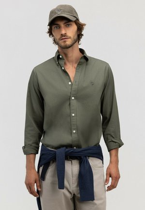 Hombre con camisa color verde oliva abotonada, mangas remangadas, pantalones claros, suéter azul marino atado a la cintura y gorra verde oliva, de pie frente a un fondo liso.