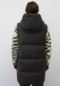 Svart pufferväst med hög krage, med en kviltad design och en slät yta. Bärs över en mönstrad tröja.