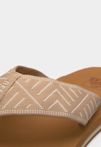 Μπεζ σανδάλι τύπου φλιπ-φλοπ με λευκό σχέδιο ζιγκ-ζαγκ στο υφασμάτινο λουράκι και ταμπά σόλα.