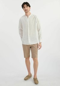 Lekka lniana koszula w kolorze białym, z button-down kołnierzykiem, zestawiona z beżowymi szortami. Obuwie w modnym stylu wsuwanym w dopasowanym odcieniu beżu.