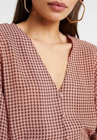 Růžová blůza s vzorem, která má výstřih ve tvaru V a houndstooth design v tmavších odstínech. Ozdobné knoflíky na přední straně. Hladká textura.