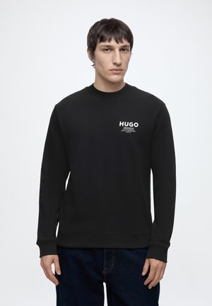 Νέος άνδρας που φοράει μαύρο φούτερ Hugo με λευκό λογότυπο στο στήθος, σε συνδυασμό με σκούρο μπλε τζιν, στέκεται μπροστά σε απλό ανοιχτό φόντο.