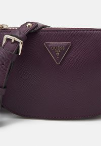 Fialová texturovaná kabelka přes rameno s kováním ve zlaté barvě a trojúhelníkovou logovou plaketou. Nastavitelný popruh s přezkou.