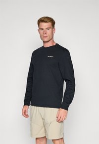 T-shirt noir en coton à manches longues avec un col rond et un logo Columbia blanc sur la poitrine, associé à un short beige avec des zippers.