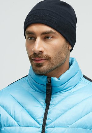 Navy fleece beanie with embossed logo, paired with a light blue quilted jacket featuring a high collar and black zipper accents.