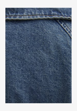 Tissu en denim avec une surface texturée, présentant des tons de bleu et des variations subtiles de couleur, avec des coutures visibles le long des bords.