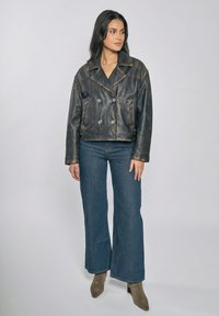 Kvinde stående iført en slidt sort læderjakke, blå vidde-bukser og taupe ankelstøvler mod en ensfarvet lys baggrund.