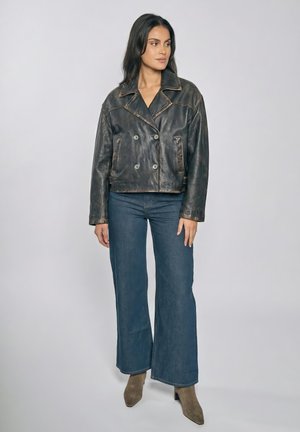 Donna in piedi che indossa una giacca di pelle nera usurata, jeans blu a gamba larga e stivaletti taupe, su uno sfondo chiaro e uniforme.