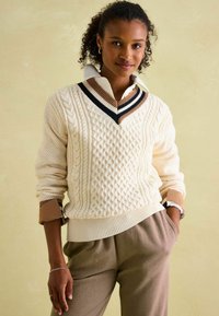 Cremefarbener Kabelstrickpullover mit V-Ausschnitt und braunen sowie navyblauen Streifenakzenten, kombiniert mit einem weißen Kragenhemd und braunen, strukturierten Hosen.