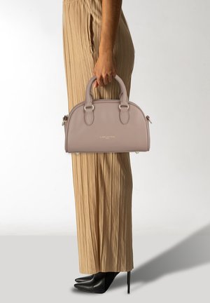 Sac à main en cuir rose clair avec un haut arrondi, deux poignées, des finitions en argent et un logo doré. Assorti à un pantalon beige plissé et des talons noirs.
