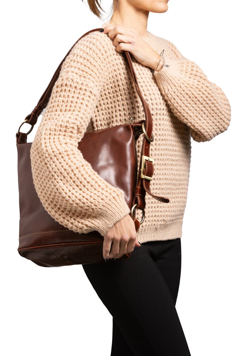 Borsa a tracolla in pelle marrone con design morbido, tracolla regolabile e accessori in metallo dorato. Indossato un maglione beige intrecciato e pantaloni neri.