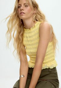 Kollane varrukateta topp, millel on kogutud tekstuur, dekoratiivsed volangid kaeluse ääres ning horisontaalsed triibud. Omadused on pehmed ja kerged.