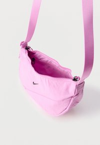 Világos rózsaszín keresztpántos táska állítható vállpánttal, fekete cipzárral és kis fekete Nike swoosh logóval az elején, fehér háttér előtt lóg.