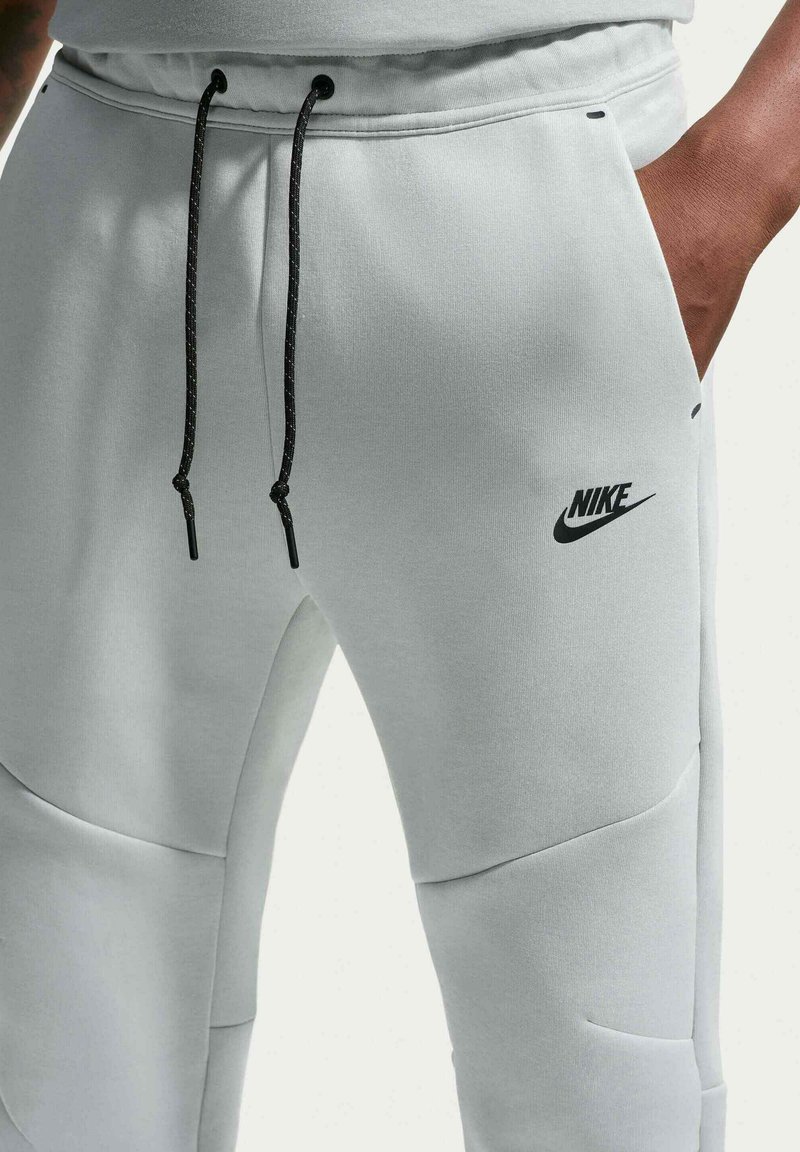 Nahaufnahme einer Person, die hellgraue Nike-Sweathosen mit schwarzen Kordeln trägt und die Hand in der Tasche hat.