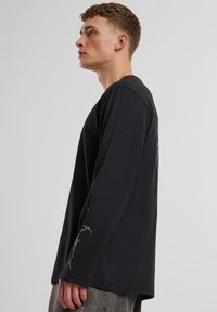 Svart langermet t-skjorte laget av bomull, med grønne grafiske design på ermene. Passformen er avslappet, og stoffet virker mykt.