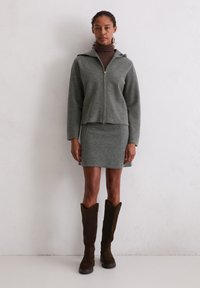 Grijze zip-up jas met capuchon, gecombineerd met een getailleerde grijze rok. Bruin suède kniehoge laarzen maken de look compleet. Gladde textuur door het geheel.