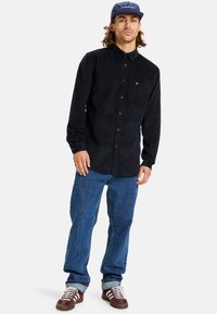 Black corduroy button-up shirt, long sleeves, single chest pocket, paired with blue denim jeans and brown sneakers with white laces.
