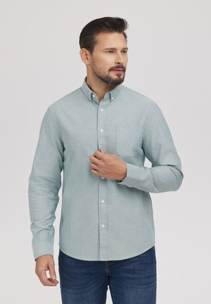 Mężczyzna w jasnoniebieskiej koszuli na guziki i niebieskich dżinsach, poprawiający koszulę prawą ręką, patrzący w prawo na jednolitym tle.