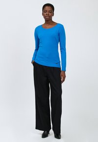 Maglione lungo blu a maniche lunghe con scollatura a barca, abbinato a pantaloni neri a gamba larga e stivali neri con tacco. Texture liscia e design aderente.