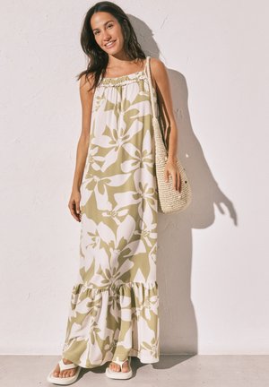 Žena se usmívá, má na sobě zelené a bílé květované maxi šaty s tenkými ramínky, drží pletenou kabelku a stojí u bílé zdi.