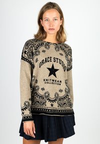 Woman with light brown hair wears beige knit sweater with black star and paisley pattern, paired with a dark pleated skirt, standing against a white background.