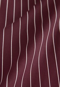 Tissu bordeaux lisse avec des fines rayures verticales blanches uniformément espacées, montrant de douces plis et une texture.
