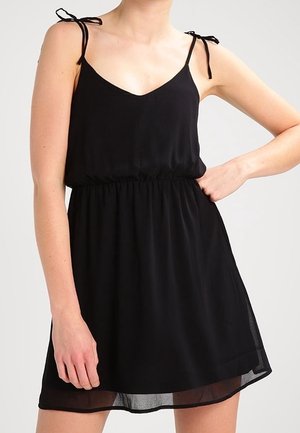 Kobieta w bezrękawną czarną sukienkę z marszczonym pasem i wiązanymi ramiączkami, stojąca z jedną ręką na biodrze na jednolitym tle.