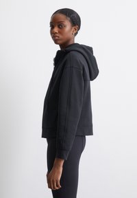 Sudadera negra con capucha, con cremallera en el frente, diseño recortado y puños acanalados. Textura suave, con costuras laterales y un ajuste relajado.