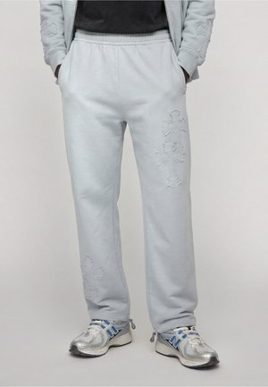 Lower body wearing light gray sweatpants with stitched cross designs and silver and blue athletic shoes, hands in pockets.