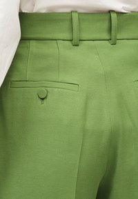 Κοντινό πλάνο πράσινων παντελονιών με ζώνη, θηλιές για ζώνη και πίσω τσέπη με κουμπί καλυμμένο με ύφασμα.