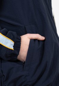 Main insérée dans la poche latérale d'une veste bleu marine foncé avec un poignet élastique rayé jaune et blanc.