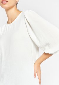 Vit blus med korta, puffade ärmar, rund halsringning, tillverkad av texturerad tyg, med en avslappnad passform och sidofickor.