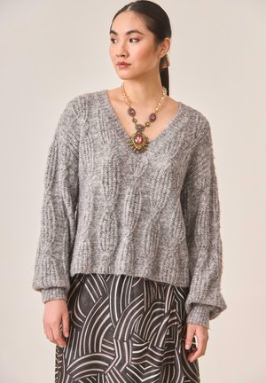 Suéter gris de punto con cuello en V, corte relajado y mangas largas. Combinado con una falda negra y plateada con estampado. Collar notable con joyas.