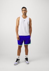 Top deportivo blanco de tirantes con textura de malla, combinado con pantalones cortos azules con detalles en verde. Zapatillas negras y blancas completan el atuendo.