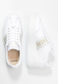 Bílé kožené tenisky s tlustými podrážkami, béžové značkové pruhy na bocích, tkaničky a "NUBIKK" vytištěné na vnitřní stélce a jazyku.