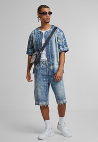 Denim outfit featuring a blue paisley print short-sleeve shirt and matching shorts. Accessories include a small black crossbody bag and sunglasses.