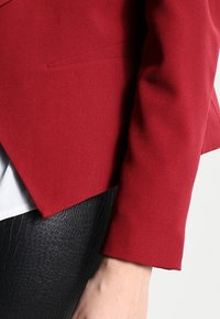 Blazer rosso su misura con una texture liscia e una forma strutturata. Presenta un bottone singolo e un'apertura frontale, abbinato a un pantalone nero testurizzato.