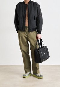 Veste bomber noire en tissu texturé, pull ras du cou marron, pantalon vert olive, baskets grises et vertes, sac fourre-tout noir avec des poignées logo.