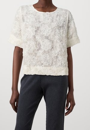 Zwart persoon met een wit topje met korte mouwen met gestructureerde bloemmotieven en donker geribde broek, staand voor een effen achtergrond.