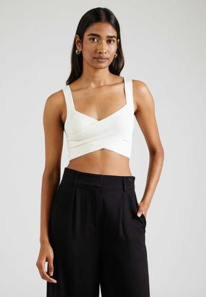 Woman wearing a white sleeveless crop top and high-waisted black wide-leg pants, standing with one hand in pocket against plain background.