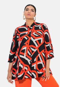 Schwarze Bluse mit Knöpfen, die auffällige rot-graue abstrakte Muster aufweist, Dreiviertelärmel und einen lässigen Schnitt hat. Kombiniert mit orangefarbenen Hosen.